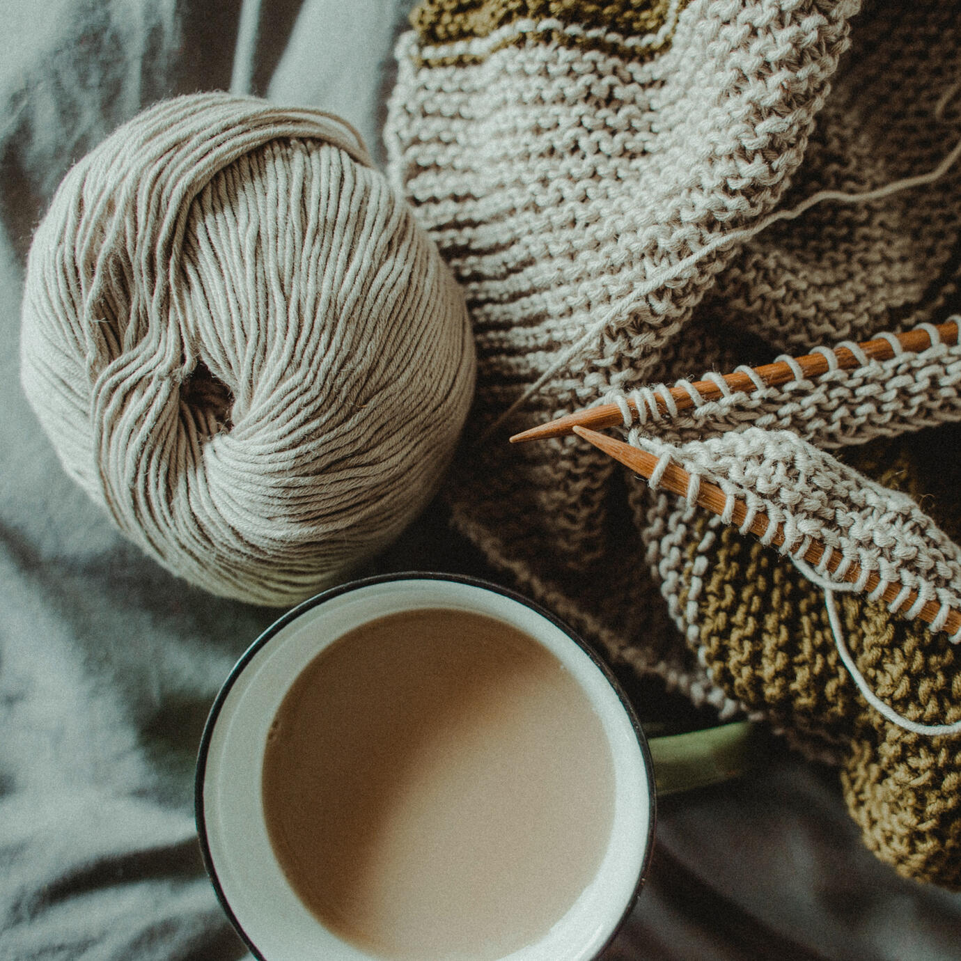 A cup of coffee sits next to a ball of white yarn, linked to two wood knitting needles. The needles are being used to create a project in garter stitch.